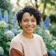 A smiling young woman with short curly dark hair, looking confident and reassuring in an outdoor setting.