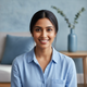 Close-up portrait of a smiling South Asian woman with dark hair, looking at the camera in a blurred blue home interior.